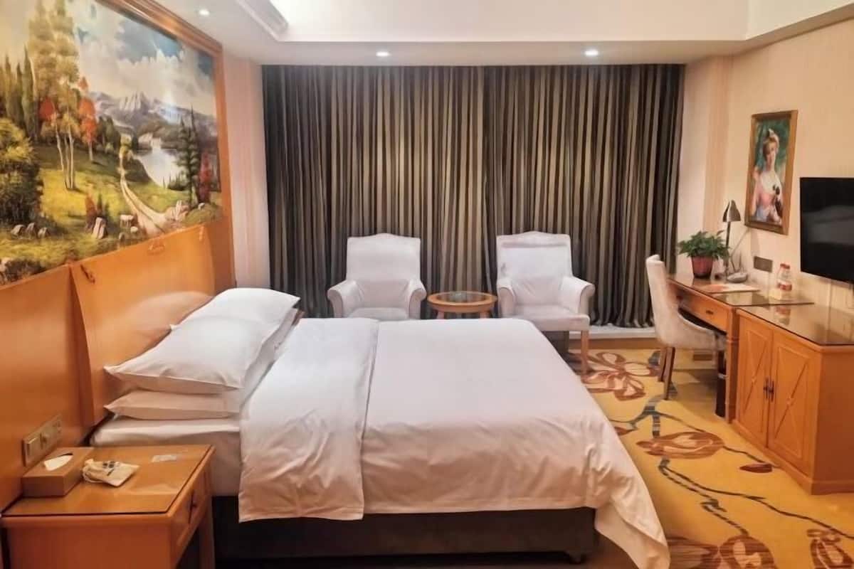 A hotel room with a bed, two chairs, a desk, a television, and a painting on the wall.