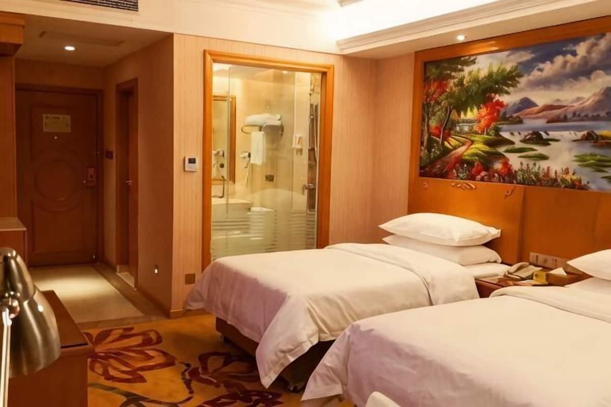A hotel room with two beds, a painting on the wall, a bathroom with a glass door, and a carpeted floor.