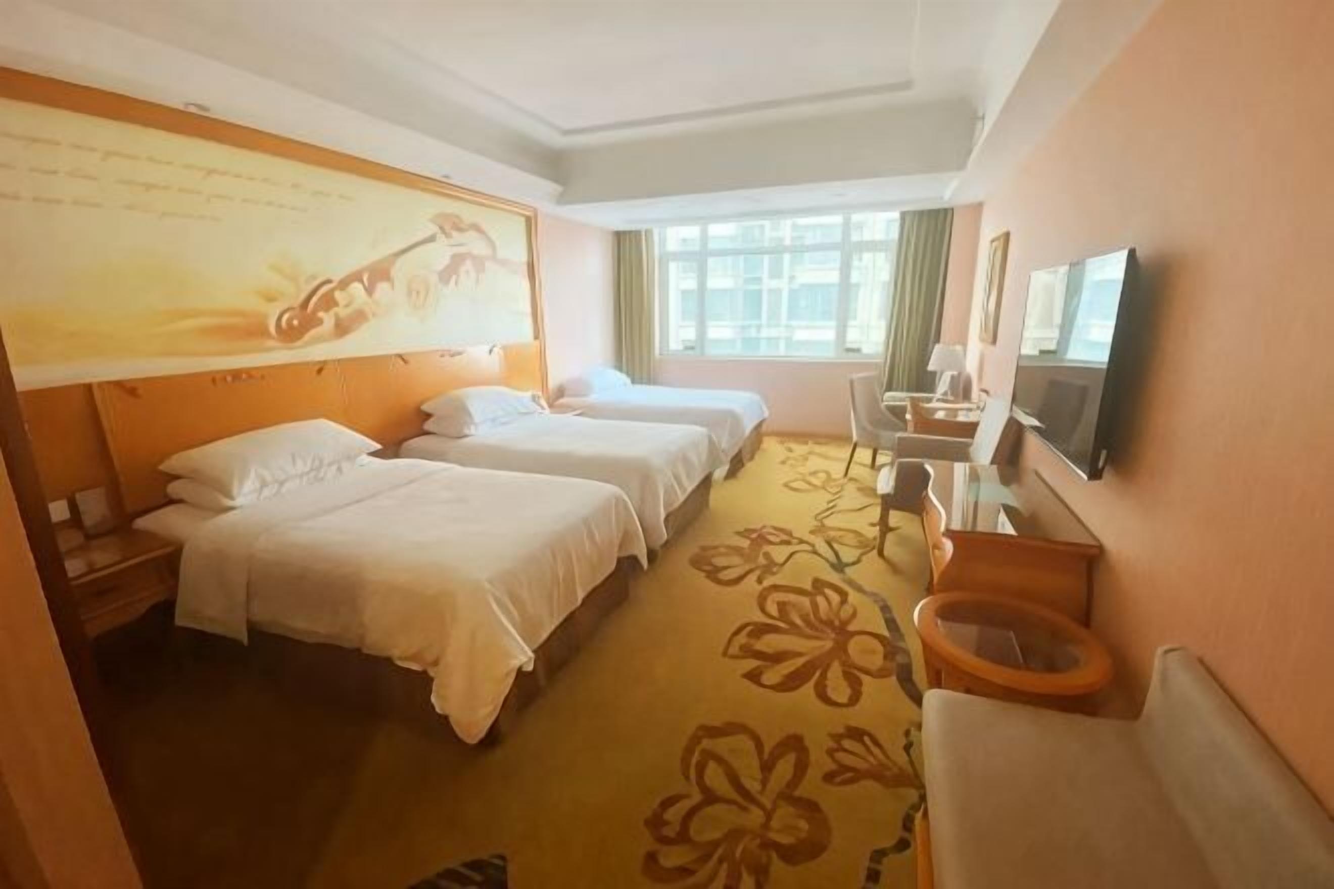 A hotel room with three beds, a television, a bathroom with a toilet, and a window with curtains.