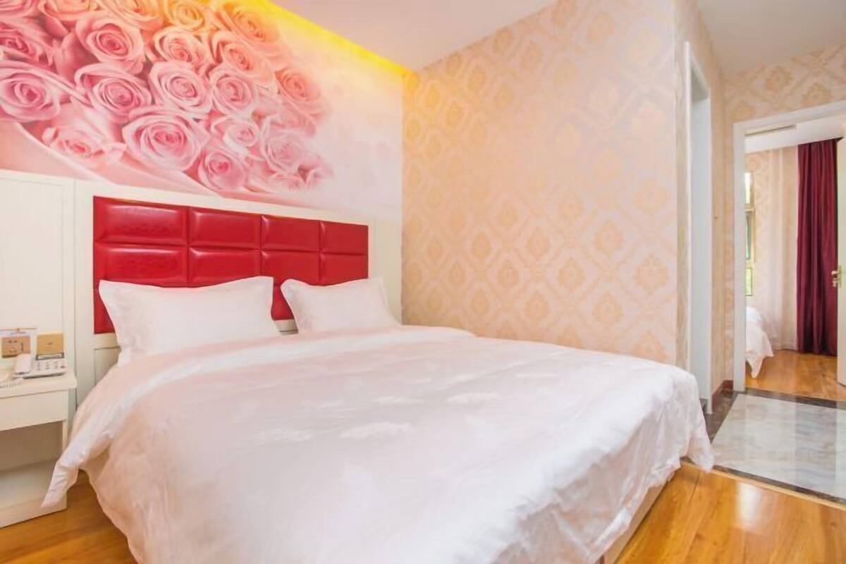 A hotel room with a bed, a nightstand, a red headboard, and a wall with a rose pattern.