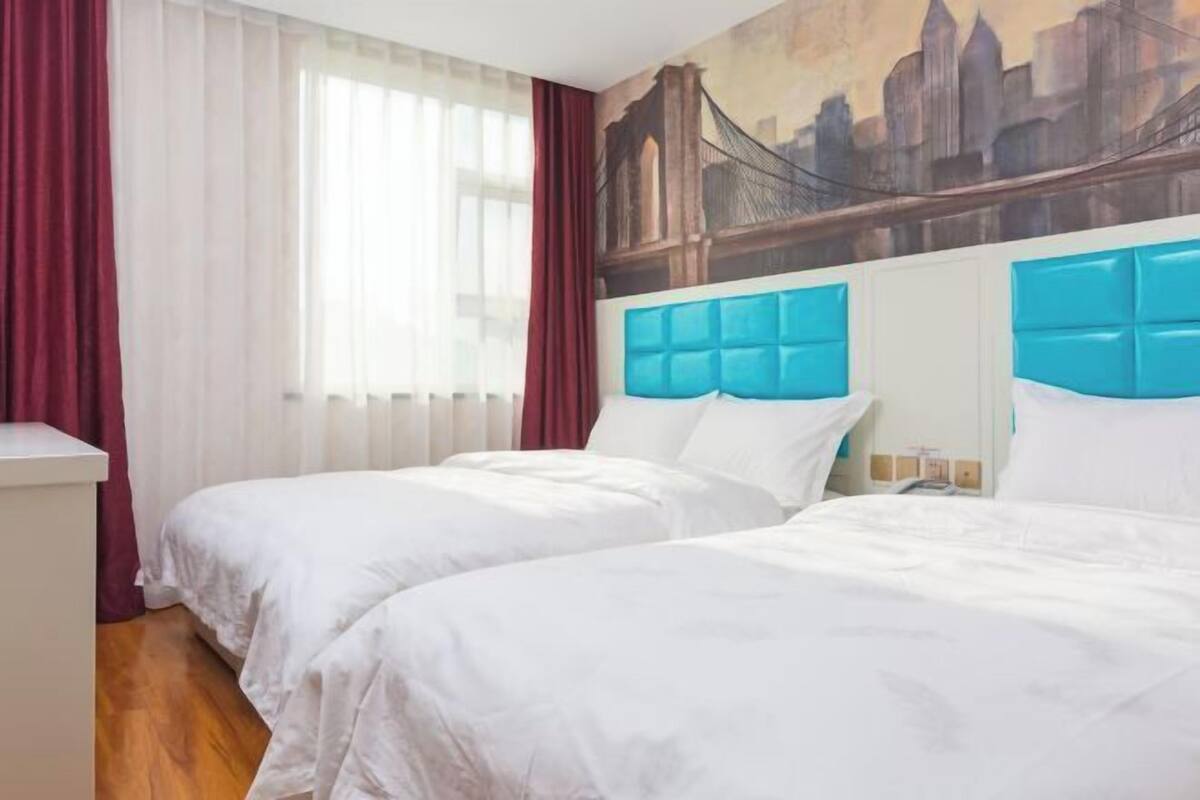A hotel room with two beds, a cityscape mural on the wall, and a window with curtains.