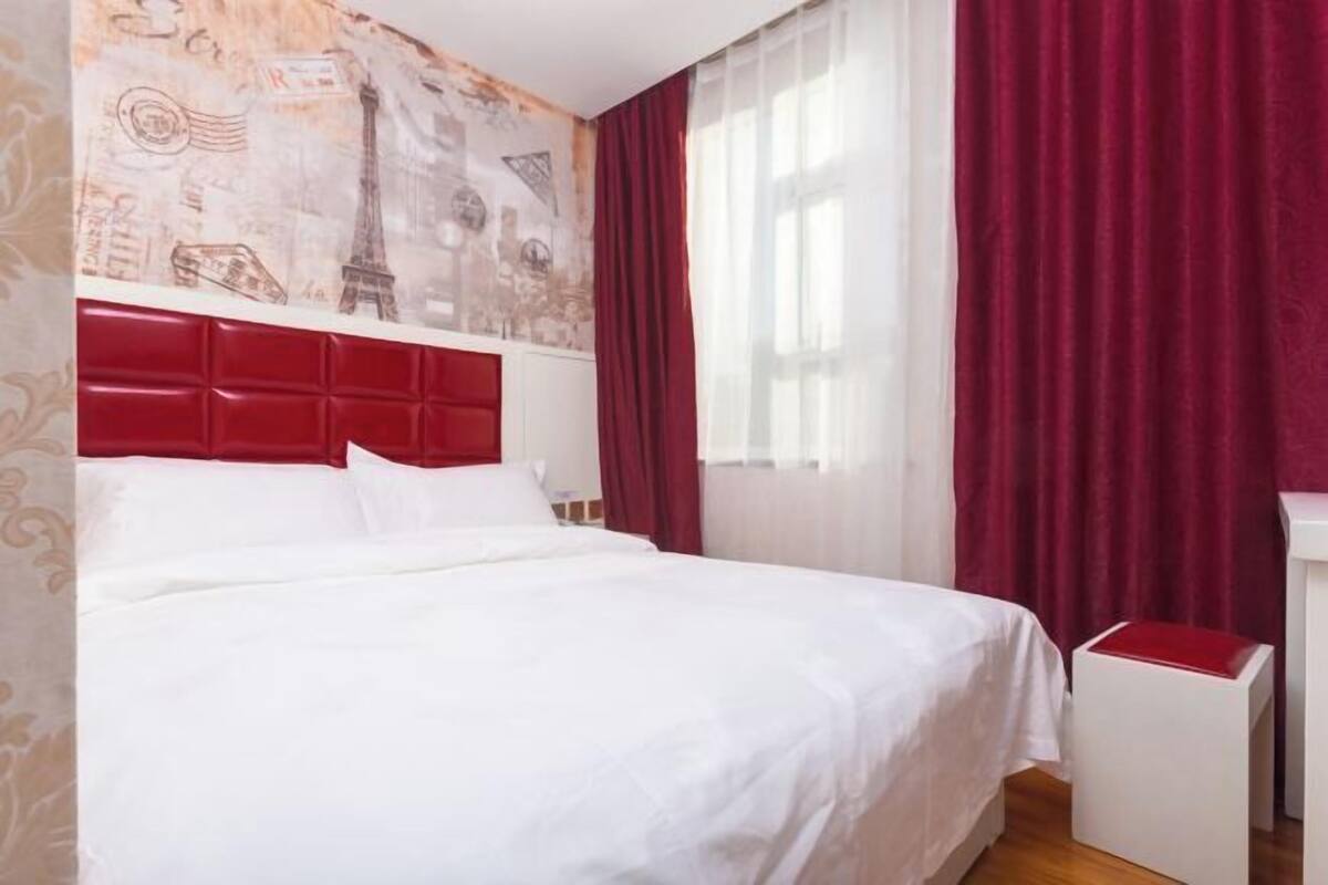 A hotel room with a bed, a red headboard, a window with curtains, and a small white table.