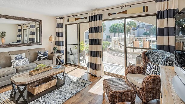 Ein geräumiges Wohnzimmer mit einer Couch, Couchtisch und Korbstühlen, großen Fenstern mit gestreiften Vorhängen und Blick auf die Stadt.
