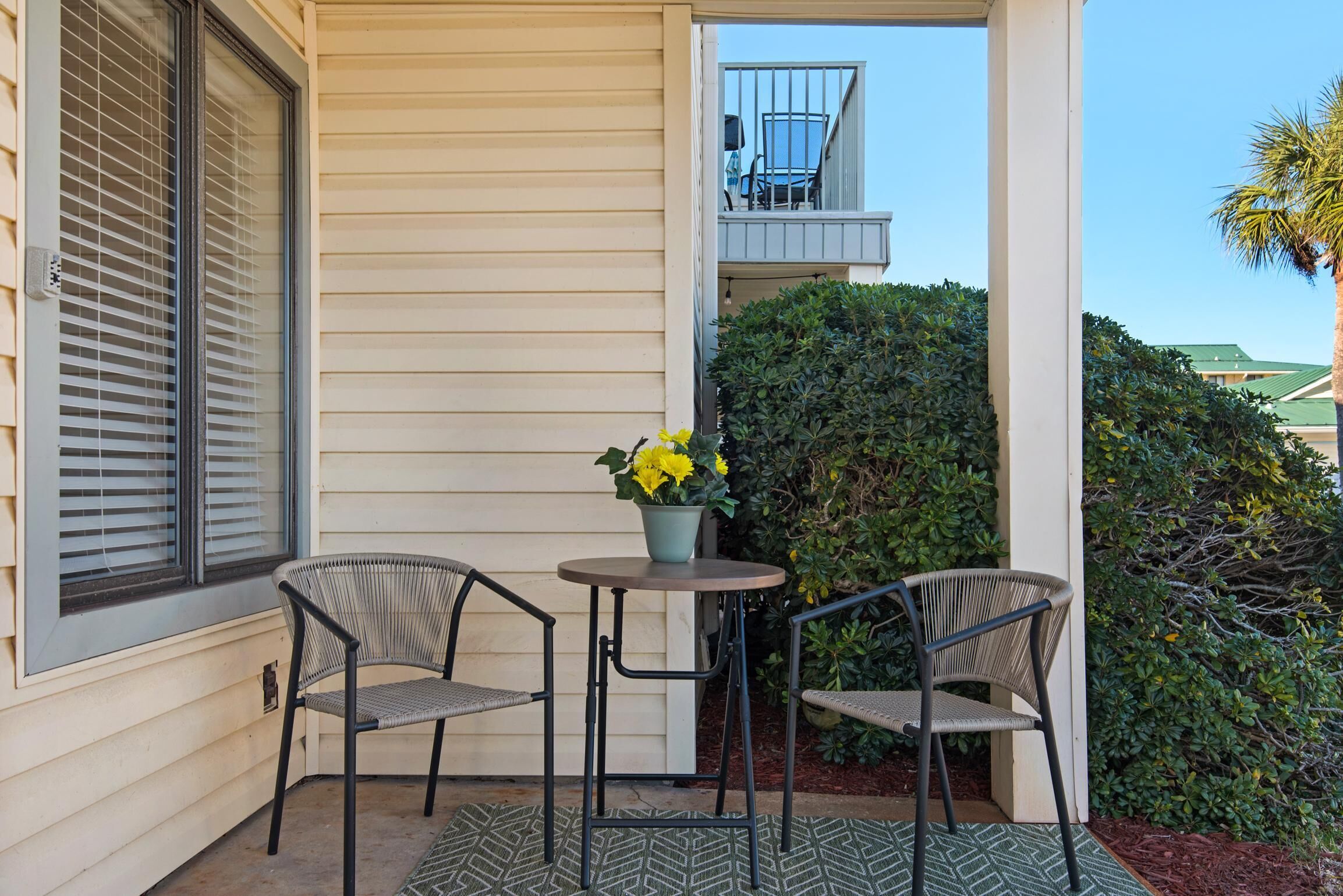 A small outdoor seating area with two chairs and a table, a potted plant with yellow flowers, and a view of a balcony with a chair.