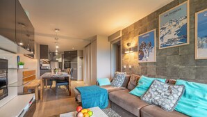 A modern living room with a brown sofa, a dining table, and a coffee table with fruit. There are framed pictures on the wall and a kitchen in the background. - Tikal  (Les Belleville)