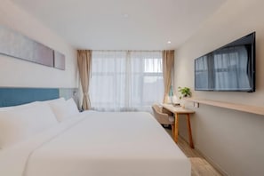 A hotel room with a large bed, a desk, a chair, a TV mounted on the wall, and a window with sheer curtains.