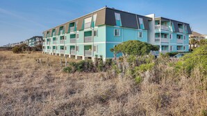 A multi-story residential building with balconies, located in a grassy area with sparse vegetation. - Balcony w/ Ocean Views: Carolina Beach Condo! (Carolina Beach)