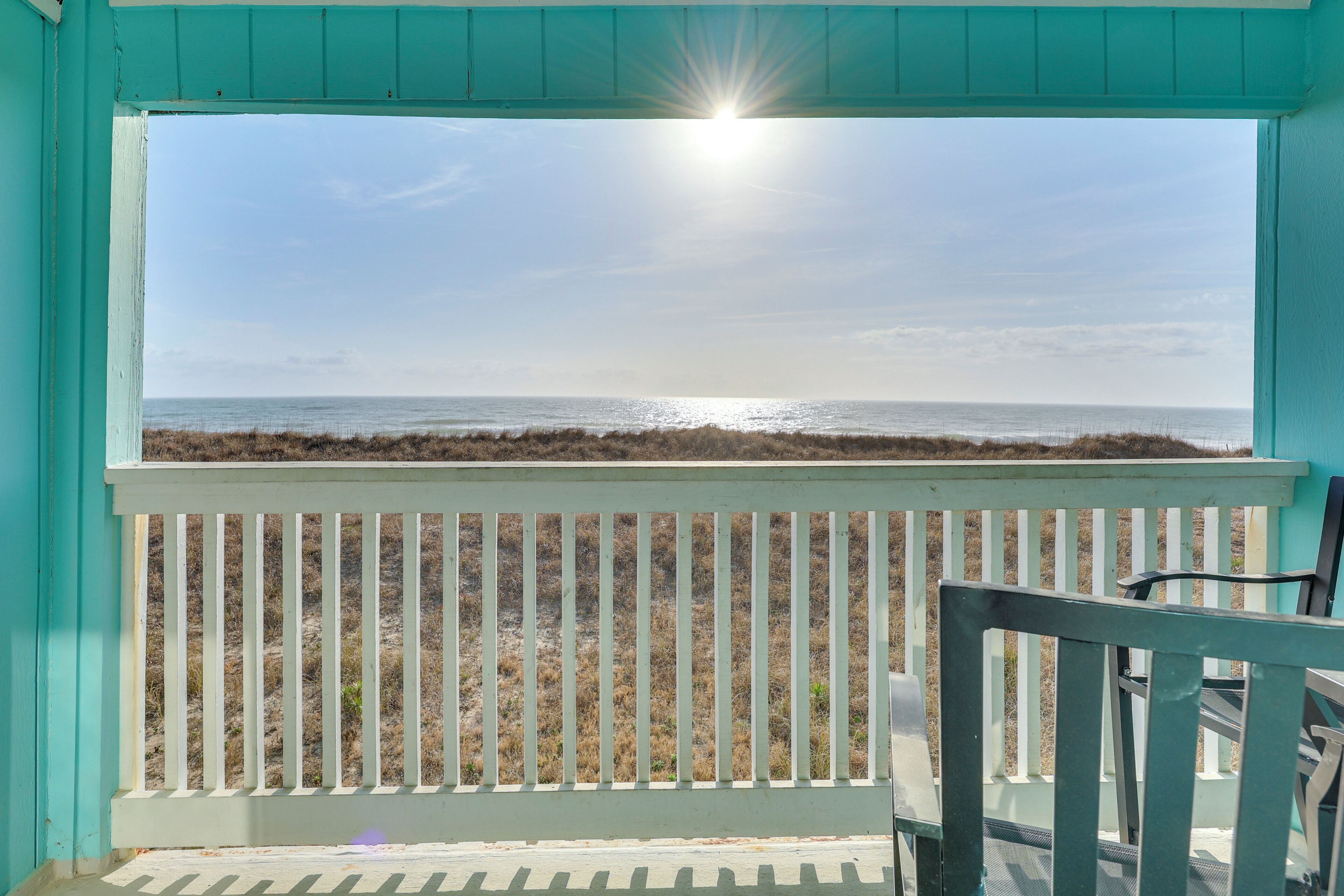 A view from a balcony with a white railing, overlooking a beach with dry grass and the ocean.
