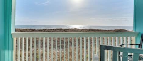 Vista desde un balcón con barandilla blanca, que da a una playa con hierba seca y el océano.