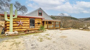 A rustic log cabin with a prominent cactus, a wooden porch, and a gravel driveway. - Fire Pit, Views: Peaceful Texas Hill Country Cabin (Tarpley)