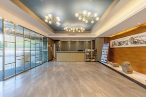 A hotel lobby with a reception desk, a large glass partition, and a wooden reception desk with a framed artwork. - Shiji Xinghe Hotel (Huangshan Tunxi Old Street) (Huangshan City)