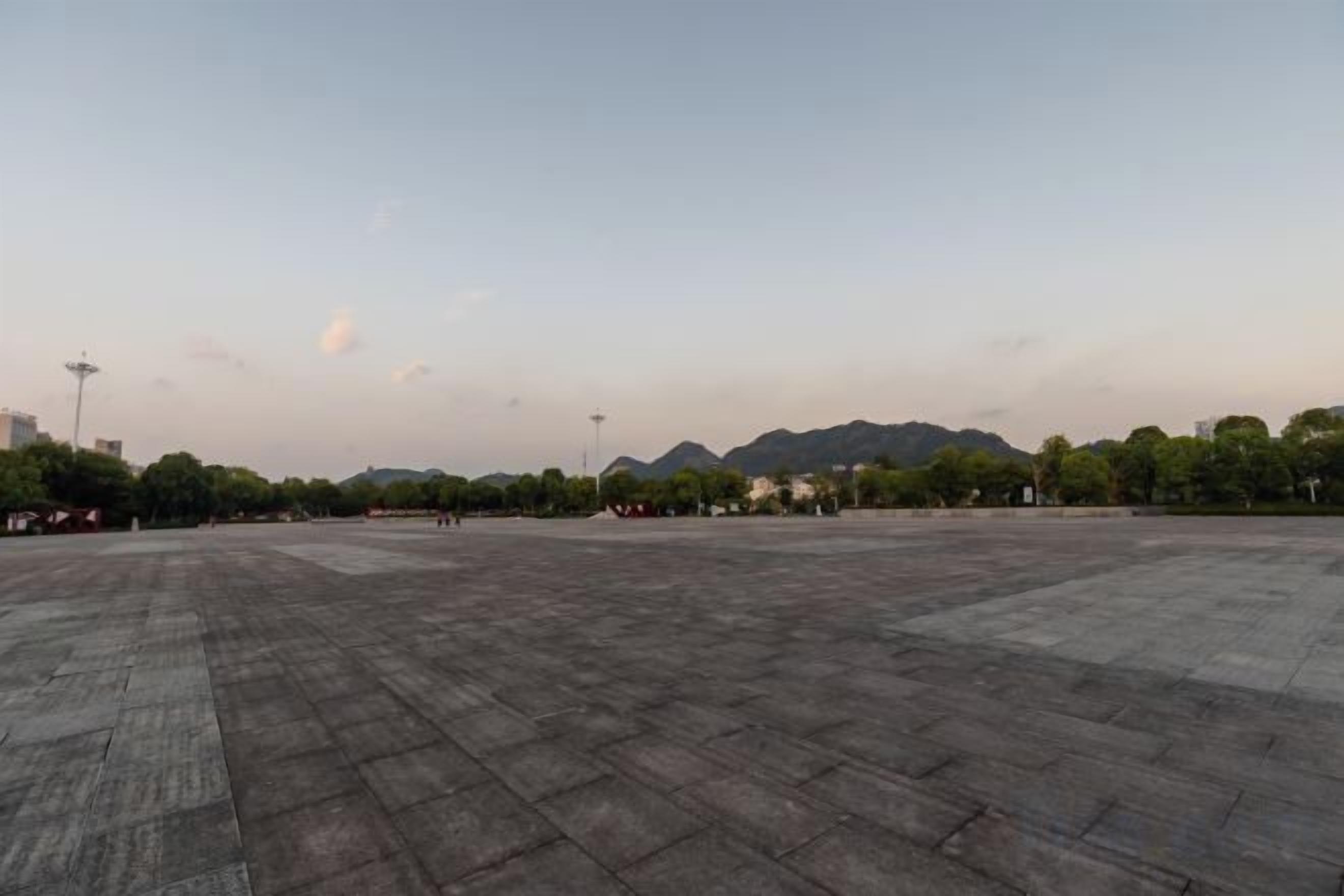 An open plaza with a paved ground, surrounded by trees and distant mountains.