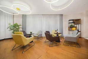 Quarto de hotel moderno com piso de madeira, uma janela grande com cortinas e um espaço de estar com cadeiras e uma mesa pequena.