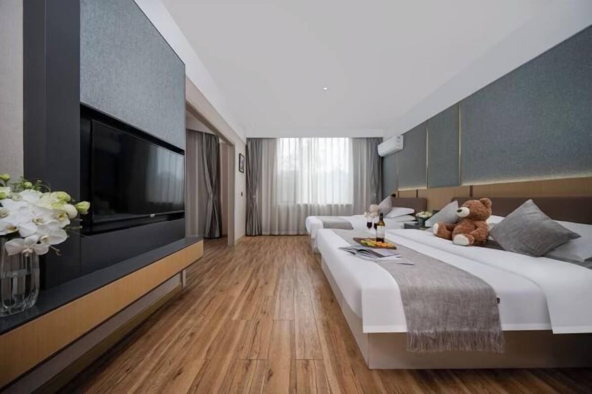 Une chambre d’hôtel moderne dotée d’un grand téléviseur à écran plat, d’un lit recouvert d’une couette blanche, d’un ours en peluche et d’un vase rempli de fleurs.