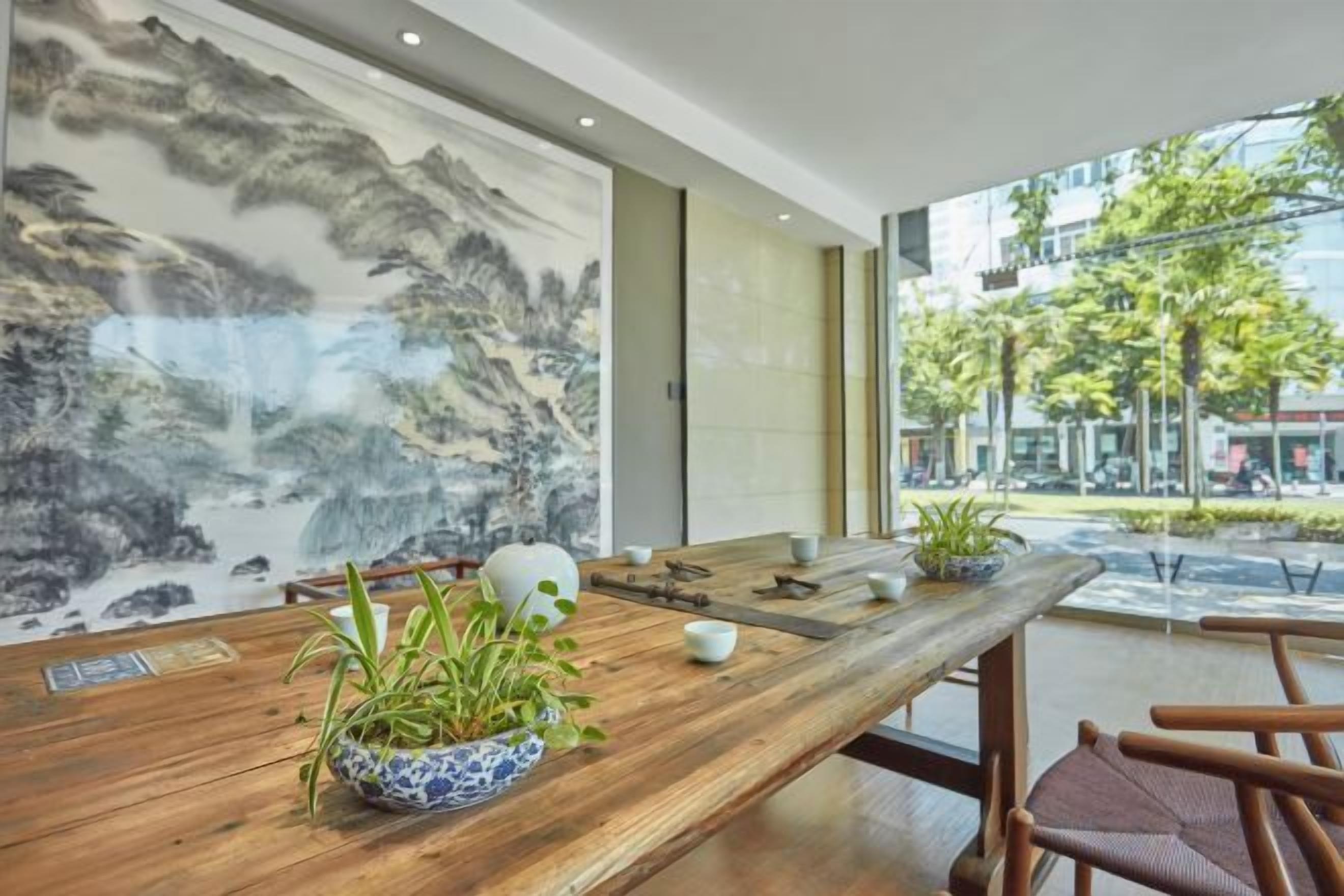 A wooden table with a bowl of plants, a white vase, and a teapot in a modern interior with large windows and a view of the street.