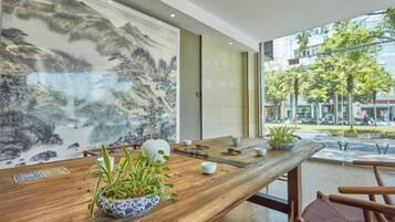 A wooden table with a bowl of plants, a white vase, and a teapot in a modern interior with large windows and a view of the street.