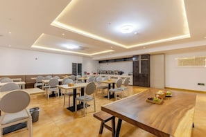 A modern dining area with tables and chairs, a kitchen in the background, and a wooden table with fruits. - Shiji Xinghe Hotel (Huangshan Tunxi Old Street) (Huangshan City)