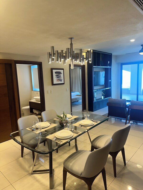 A modern dining area with a glass table, four chairs, and a contemporary pendant light fixture.