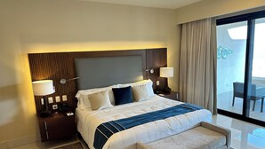 Habitación de hotel con una cama grande, dos mesitas de noche, un banco, vista al balcón y una ventana con cortinas.