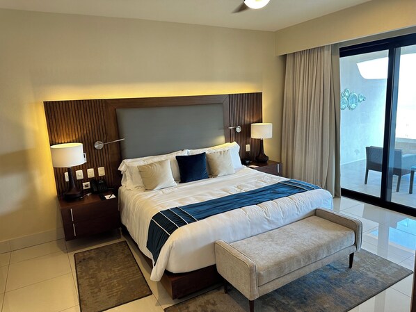 Habitación de hotel con una cama grande, dos mesitas de noche, un banco, vista al balcón y una ventana con cortinas.