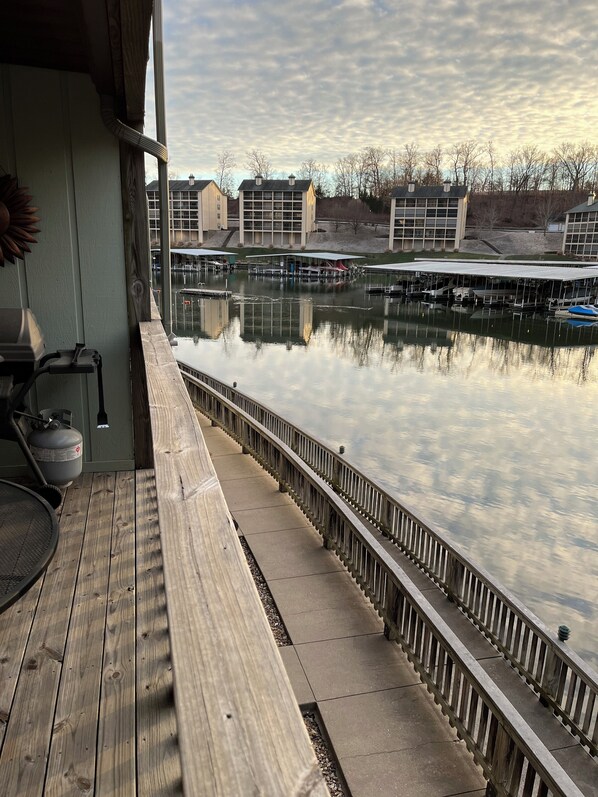 Una terraza de madera con barandilla da vista a un cuerpo de agua tranquilo, muelles y edificios al fondo.