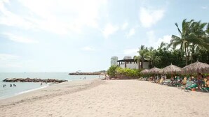 Una playa con sombrillas de paja, palmeras y un edificio al fondo.