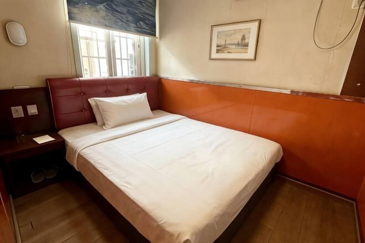 A hotel room with a bed, a nightstand, a window, and a painting on the wall.