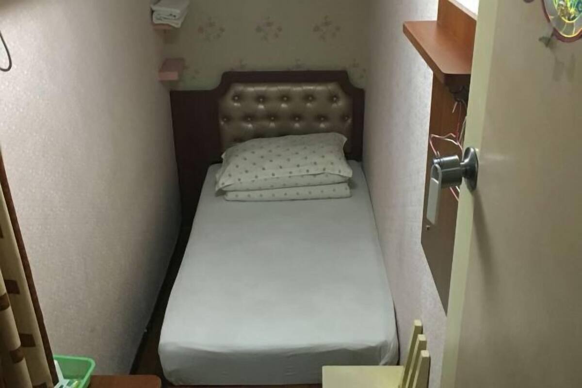 A small, narrow room with a single bed, a wooden headboard, and a door with a handle.