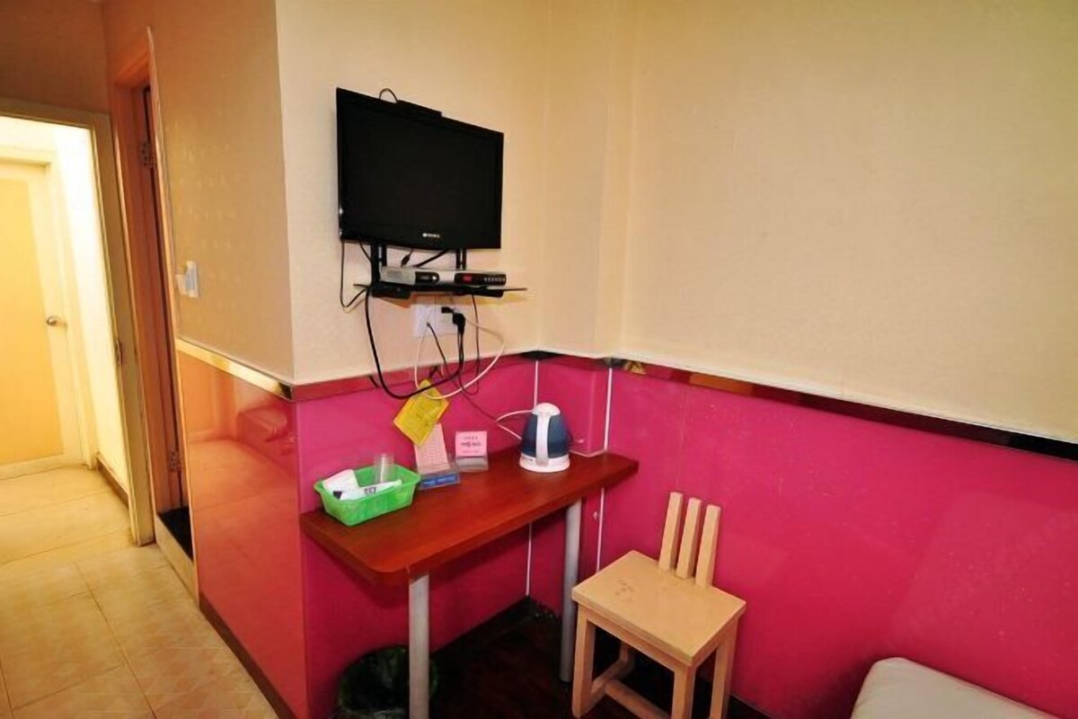 A room with a television mounted on the wall, a small wooden table with a chair, and a pink wall.