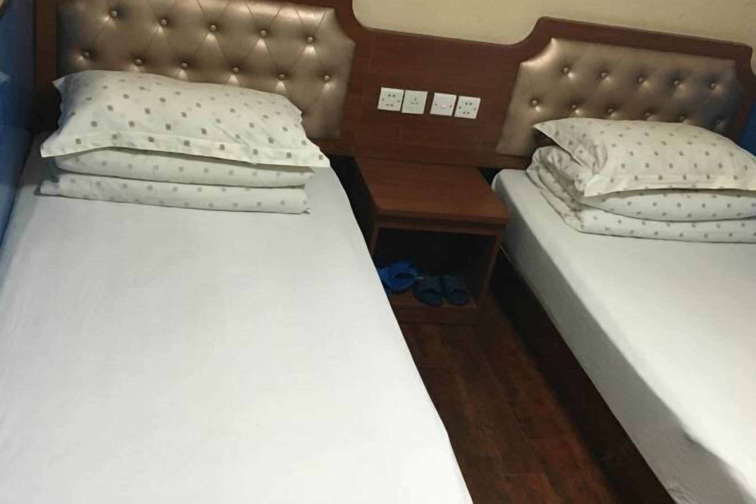 Uma cama de casal com cabeceira estofada, um criado-mudo de madeira e um par de chinelos azuis no chão.