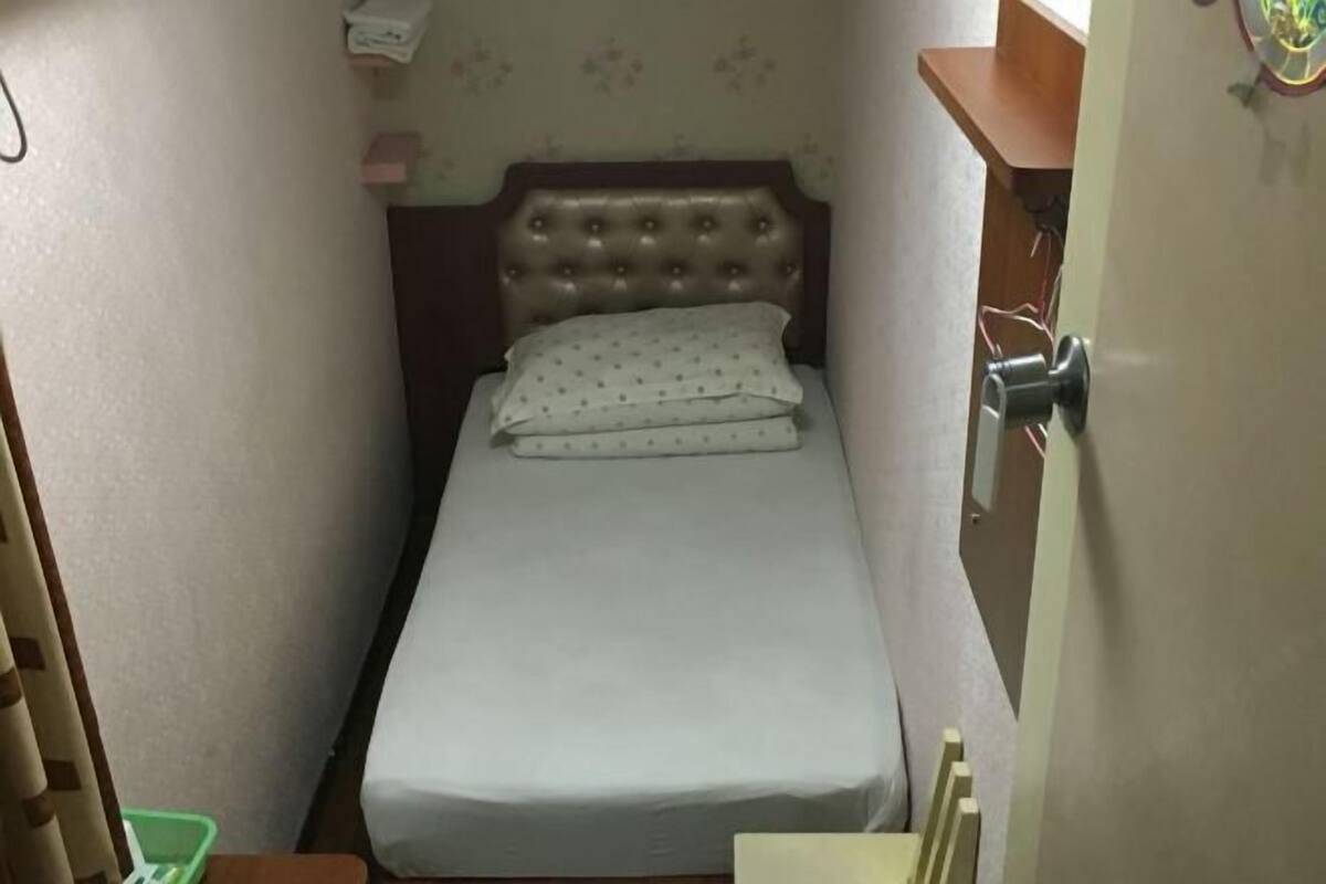 A small, single-bed room with a wooden headboard, a white mattress, and a green plastic container on the floor.