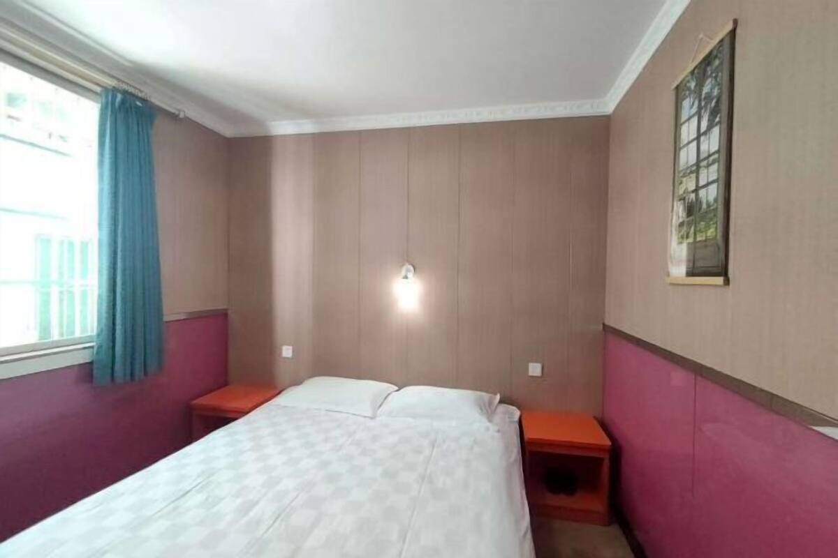 A bedroom with a bed, a nightstand, a wall-mounted light, and a window with curtains.