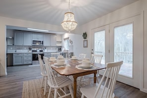 A dining area with a wooden table set for six, white chairs, a chandelier, and a kitchen in the background. - Juliet II Rental: Near Downtown & Trail Heads (Grand Junction)