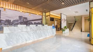 Zona de recepción de hotel con mostrador de mármol, un mural con vista a la ciudad y una escalera que sube a otro piso.