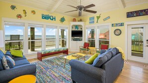A spacious living room with a large window overlooking the ocean, a ceiling fan, and a colorful rug.
