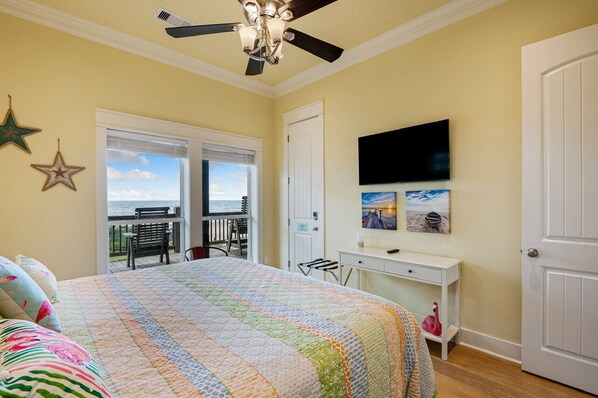 A bedroom with a large bed, a ceiling fan, a desk with a TV, and a balcony with a view of the ocean. - BEACHFRONT with DECK Hot Tub and Sunrise Views/Fishing/Steps to Beach (Bolivar Peninsula)