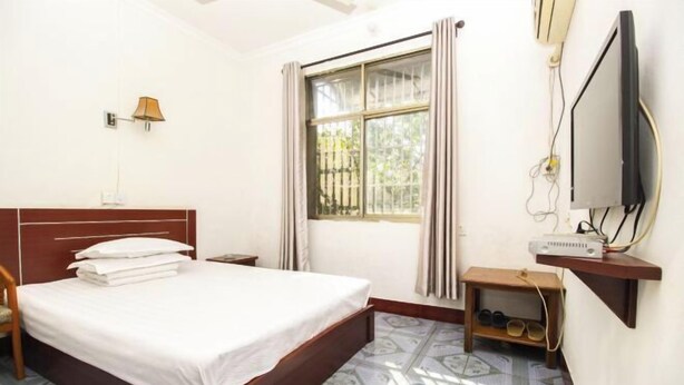 A hotel room with a bed, a TV, a desk with a computer, a window with curtains, and a shoe rack.