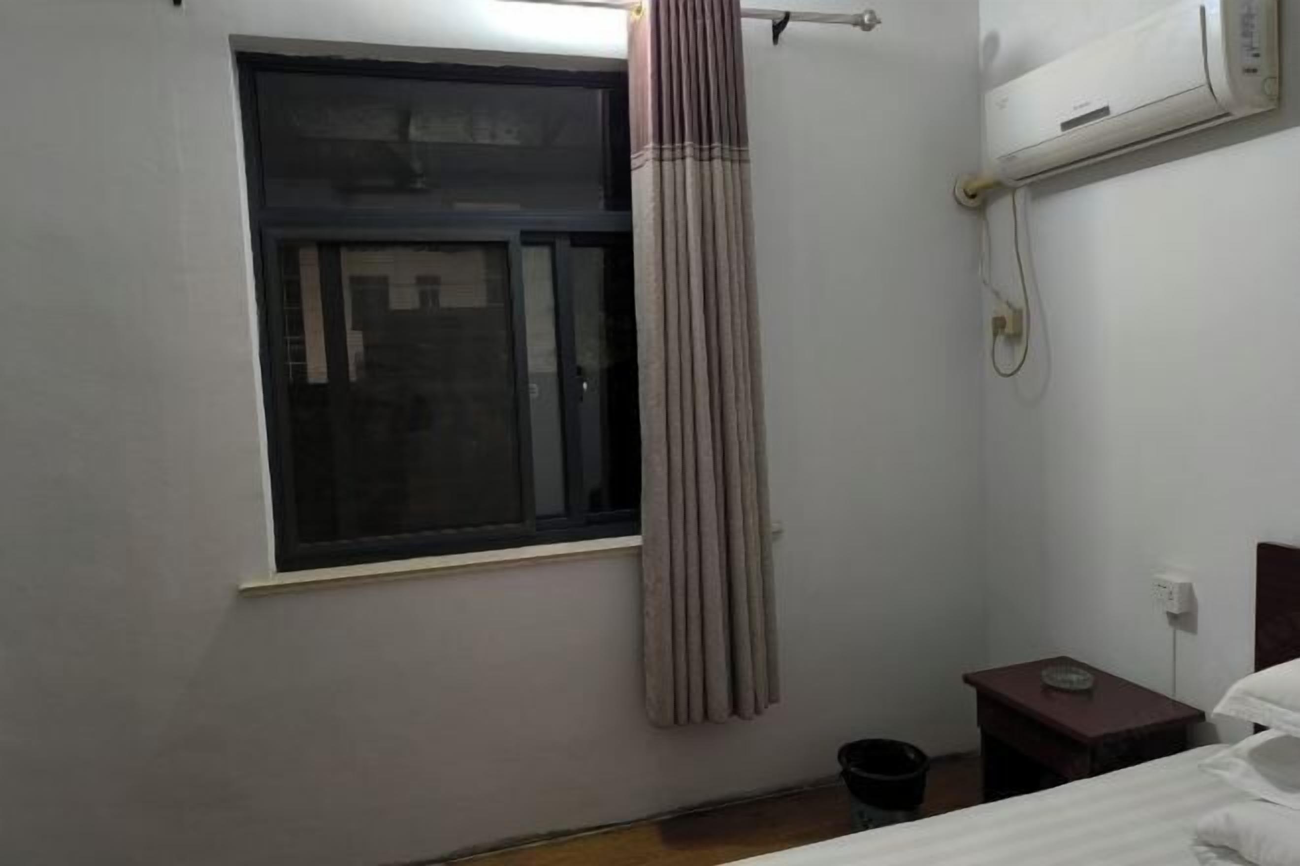 Quarto pequeno e pouco mobiliado, com uma cama de solteiro, uma mesinha, uma janela com cortina e um aparelho de ar condicionado na parede.