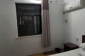 A small, sparsely furnished room with a single bed, a small table, a window with a curtain, and an air conditioning unit on the wall. - Guangde Linyuan Hotel (Guangde)