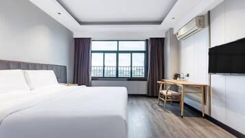 Quarto de hotel com cama, escrivaninha, cadeira, janela com cortinas e televisão na parede.