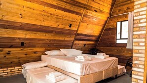 Un dormitorio acogedor en el ático con paredes de madera, techo inclinado y un ventanal grande. Hay tres camas dispuestas en forma de U, cada una con ropa de cama y almohadas impecables.