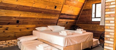Un dormitorio acogedor en el ático con paredes de madera, techo inclinado y un ventanal grande. Hay tres camas dispuestas en forma de U, cada una con ropa de cama y almohadas impecables.