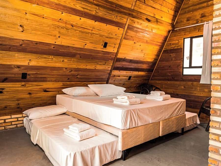 A cozy attic bedroom with wooden walls, a sloped ceiling, and a large window. There are three beds arranged in a U-shape, each with neatly made linens and pillows.