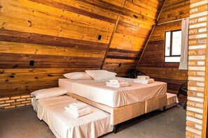 A cozy attic bedroom with wooden walls, a sloped ceiling, and a large window. There are three beds arranged in a U-shape, each with neatly made linens and pillows. - Pousada Bellíssima Itália (Luis Correia)