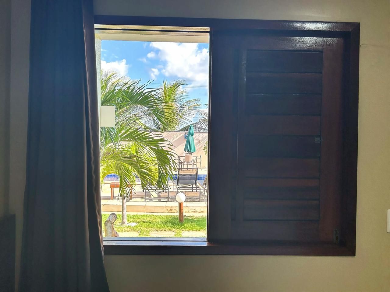 A room with a window, a wooden door, and a view of an outdoor area with a palm tree and a pool.