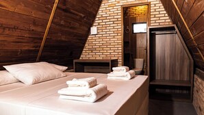 Un acogedor dormitorio en el ático con techo inclinado, pared de ladrillo y suelo de madera. Se observa una cama con ropa blanca y almohadas, una mesita de noche de madera y una puerta que da a un baño.