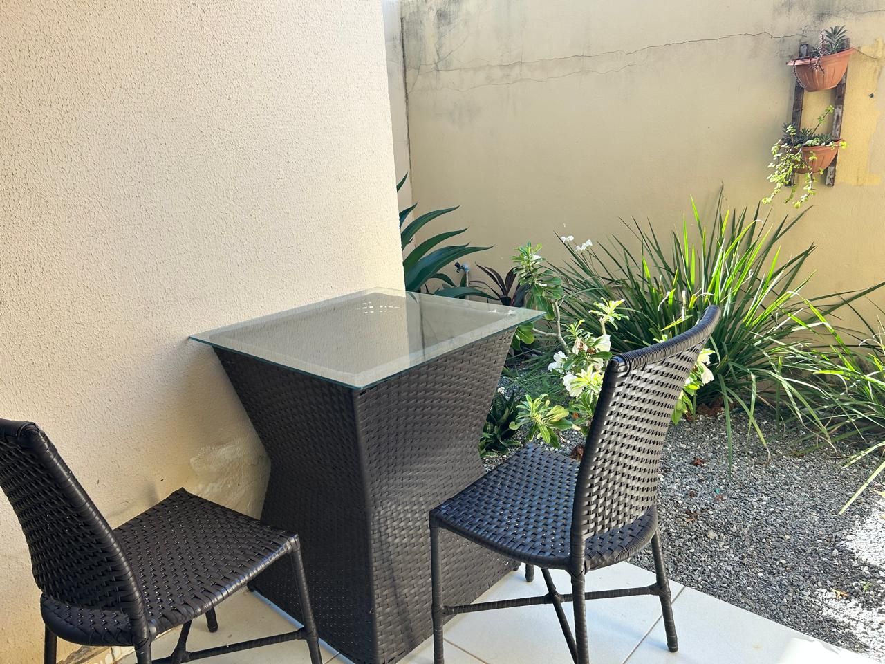 Une table à plateau en verre, accompagnée de deux chaises en osier, placée contre un mur orné de plantes en pot.