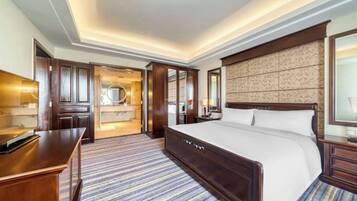Quarto de hotel com cama grande, cabeceira de madeira, uma mesa de trabalho e um banheiro visível por uma porta aberta.