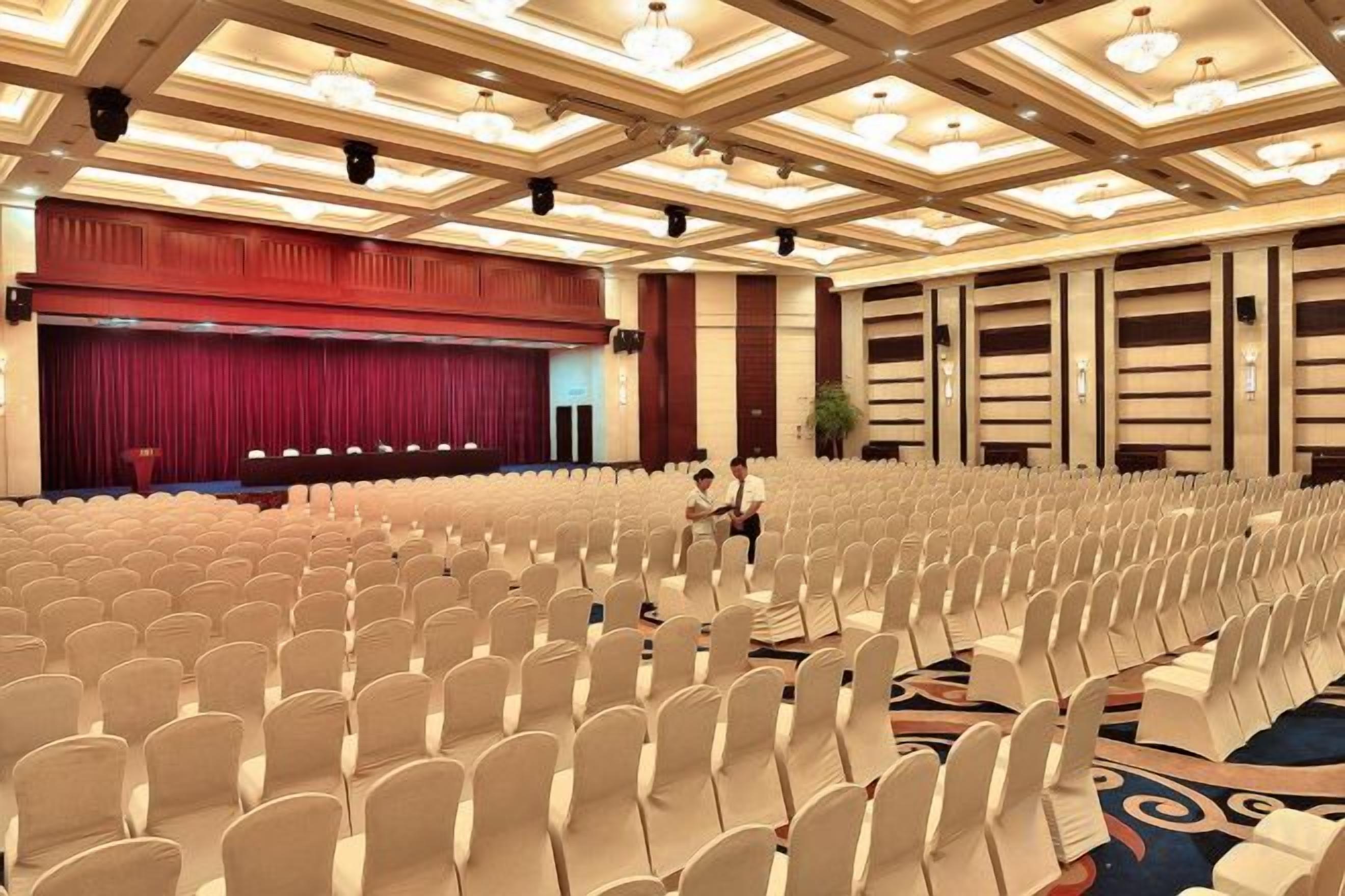 A large conference room with rows of chairs, a stage with a red curtain, and a chandelier.