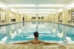 A large indoor swimming pool with a person relaxing at the edge, surrounded by chairs and a bar area.
